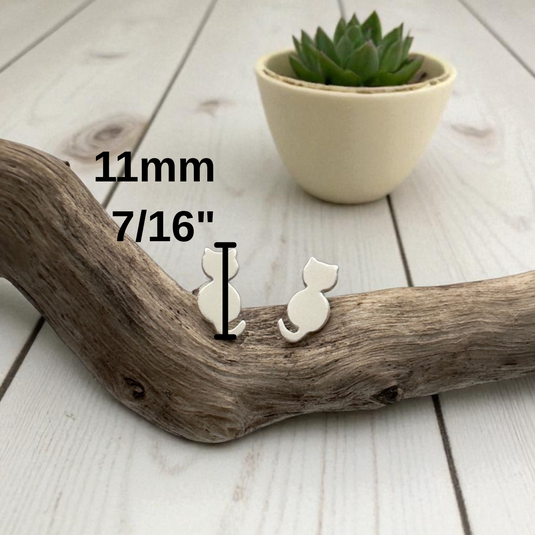 Small white cat-shaped earring on a wooden branch with measurement text and a potted plant in the background.