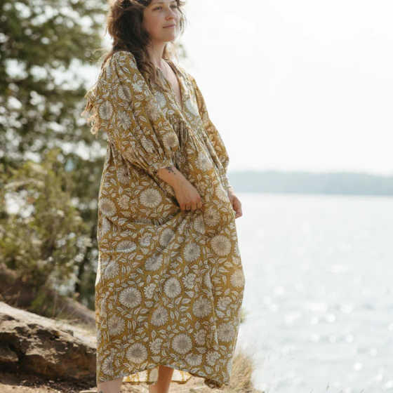 Load image into Gallery viewer, Woman in a yellow floral dress standing by a body of water with trees in the background
