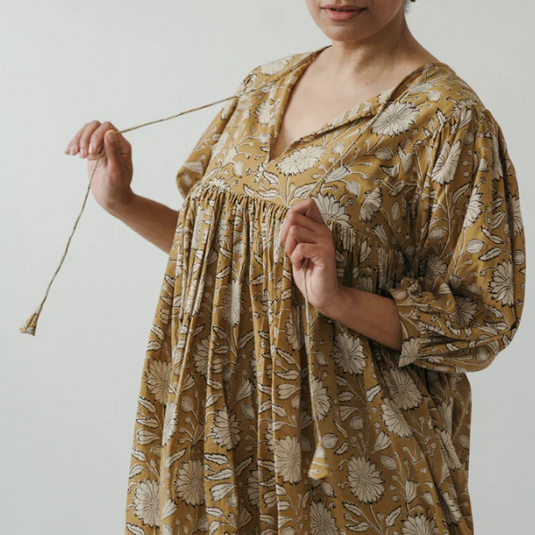 Person wearing a mustard yellow floral dress holding knitting needles against a plain background